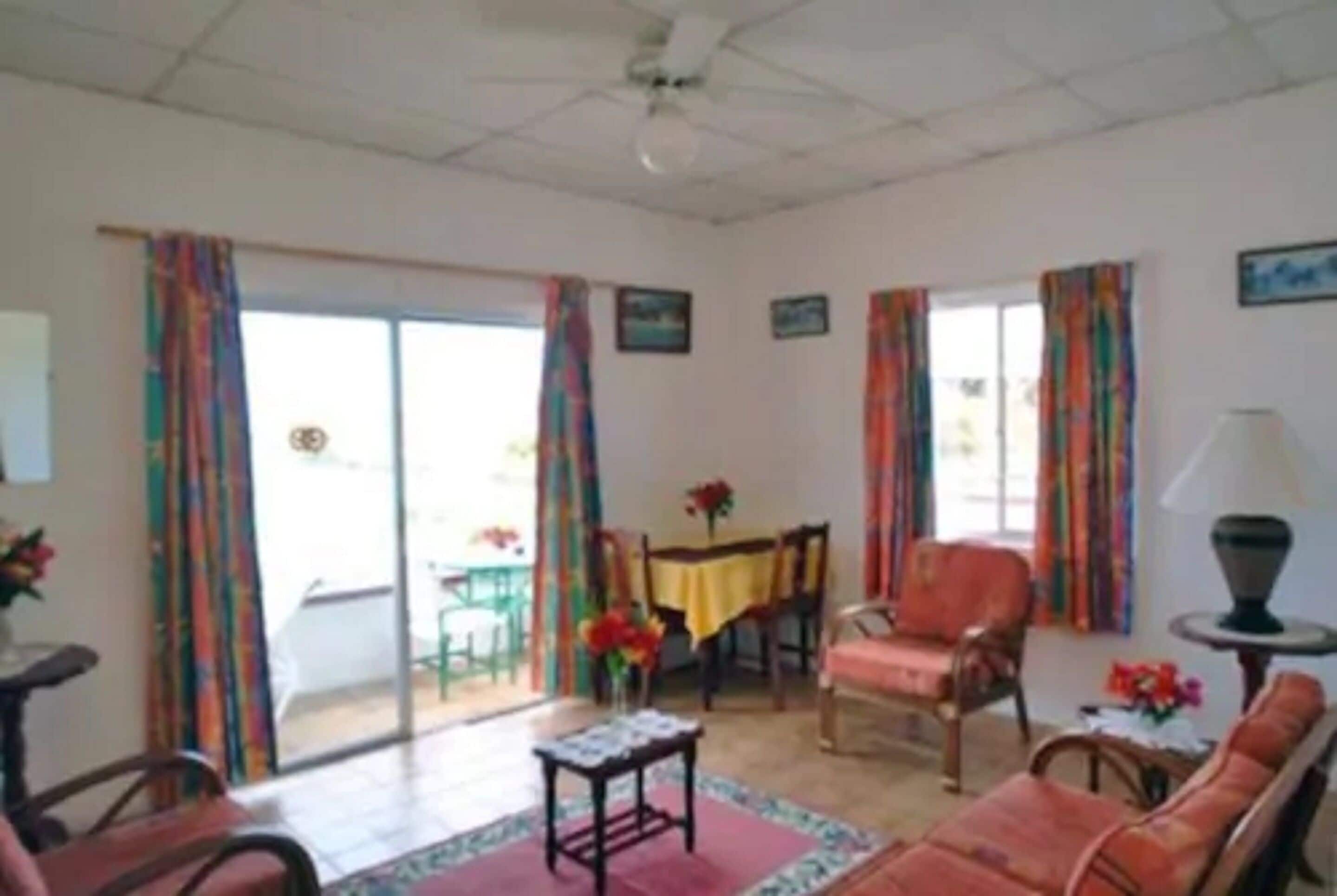 Una sala de estar con un sofá, una mesa y una silla. Hay una ventana con cortinas y una puerta que da al exterior.