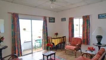 Una sala de estar con un sofá, una mesa y una silla. Hay una ventana con cortinas y una puerta que da al exterior.