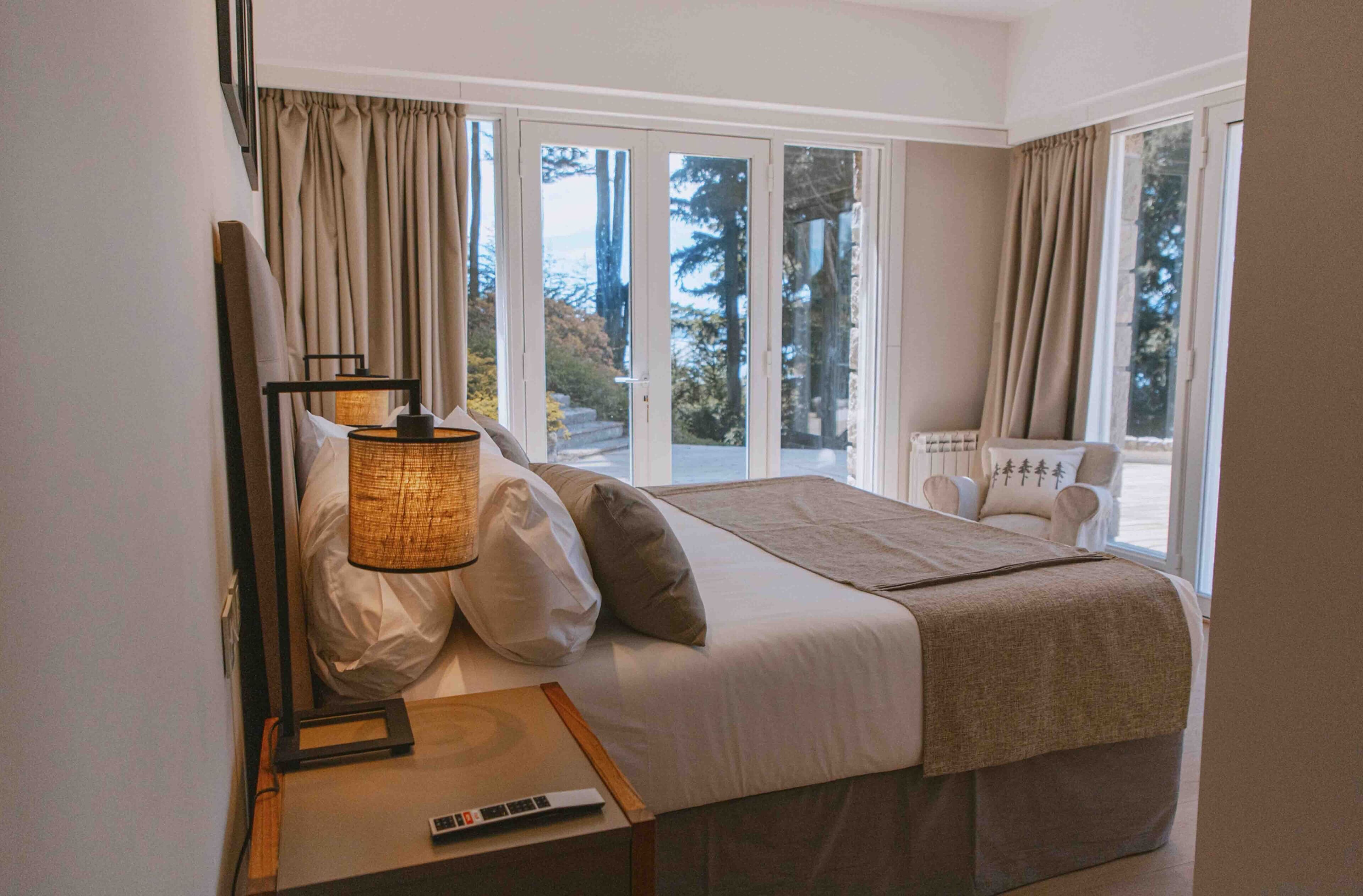 A bedroom with a bed, a bedside table with a lamp, a chair, and a view of trees through large windows.