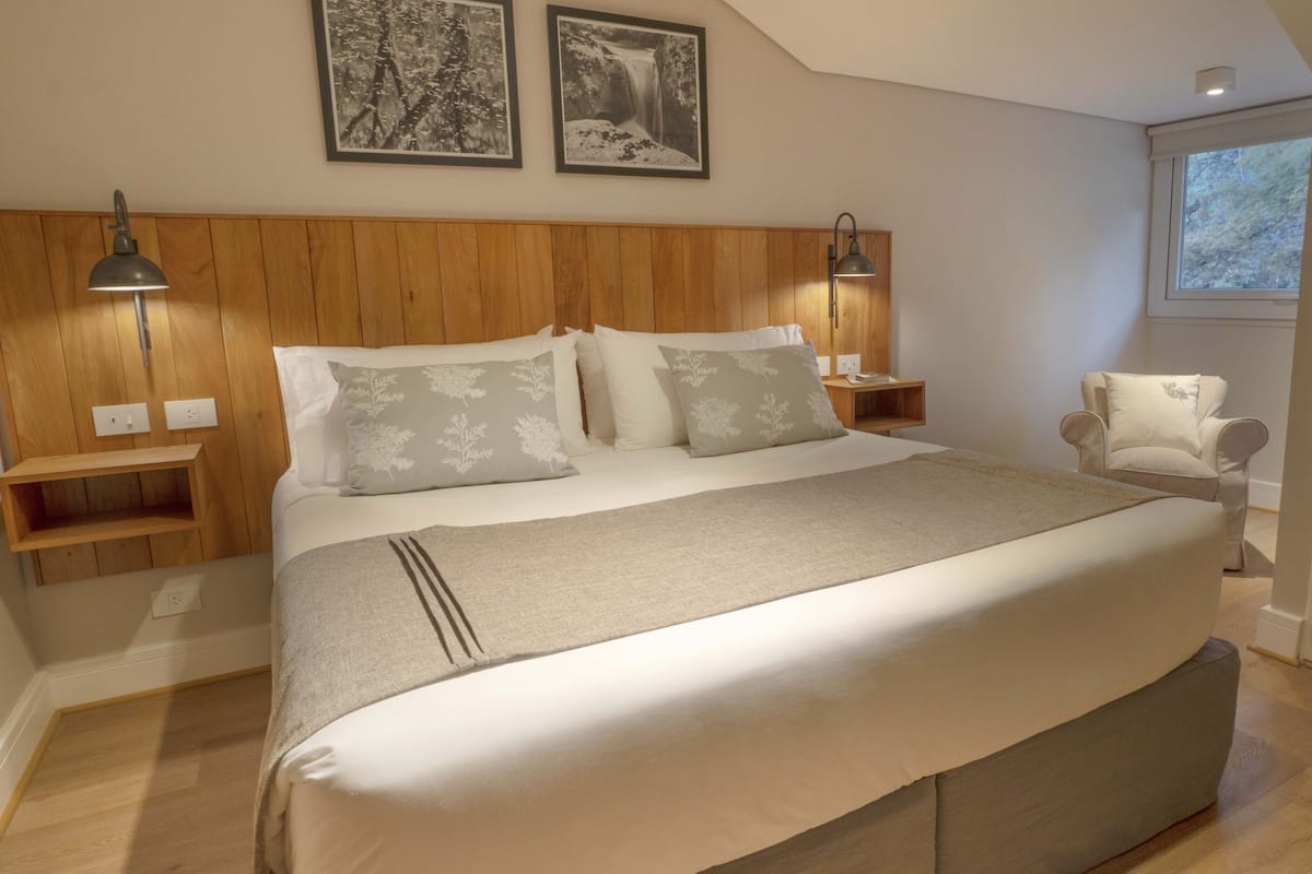 Ein Schlafzimmer mit hölzernem Kopfteil, zwei Nachttischen, einem Bett mit weißer Bettwäsche und grauen Kissen, einem Sessel und zwei Wandleuchten.