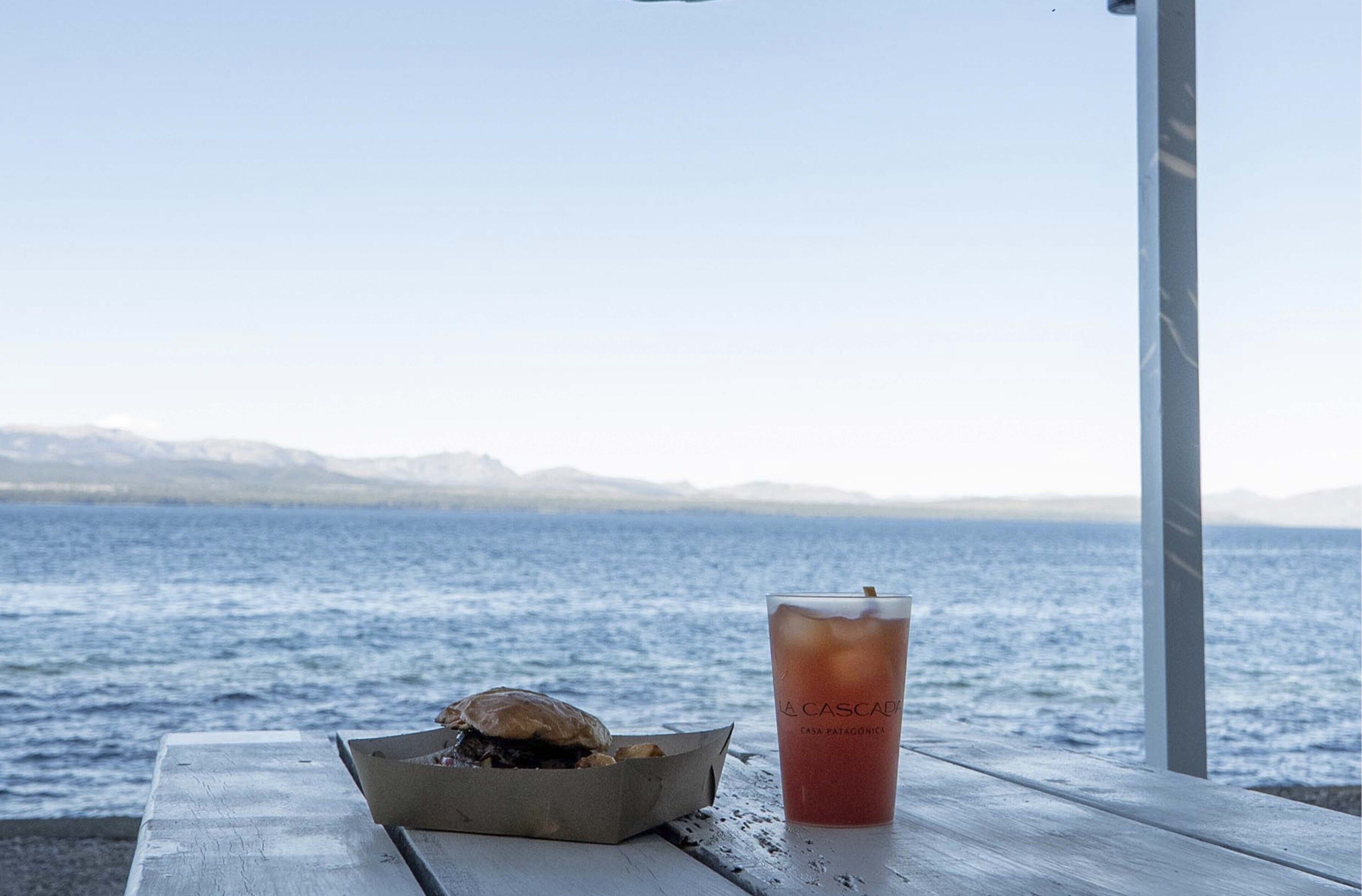 Mesa de madera con un sándwich y una bebida con el rótulo "La Cascada" de Casa Patagonia, con vistas a un cuerpo de agua y montañas distantes.