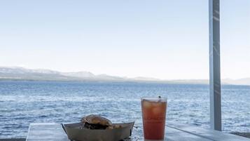 Mesa de madera con un sándwich y una bebida con el rótulo "La Cascada" de Casa Patagonia, con vistas a un cuerpo de agua y montañas distantes.