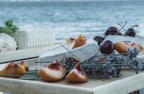 Un plateau de fromages variés et de fruits, disposés sur une planche en bois, avec en arrière-plan un plan d’eau.