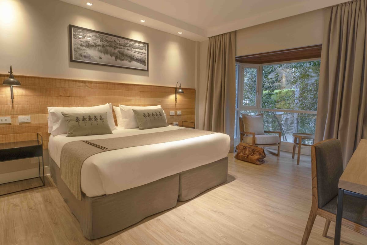 Ein modernes Hotelzimmer mit einem großen Bett, Nachttischlampen, einem hölzernen Kopfteil, einem Essbereich mit Tisch und Stühlen sowie einem großen Fenster mit Vorhängen.