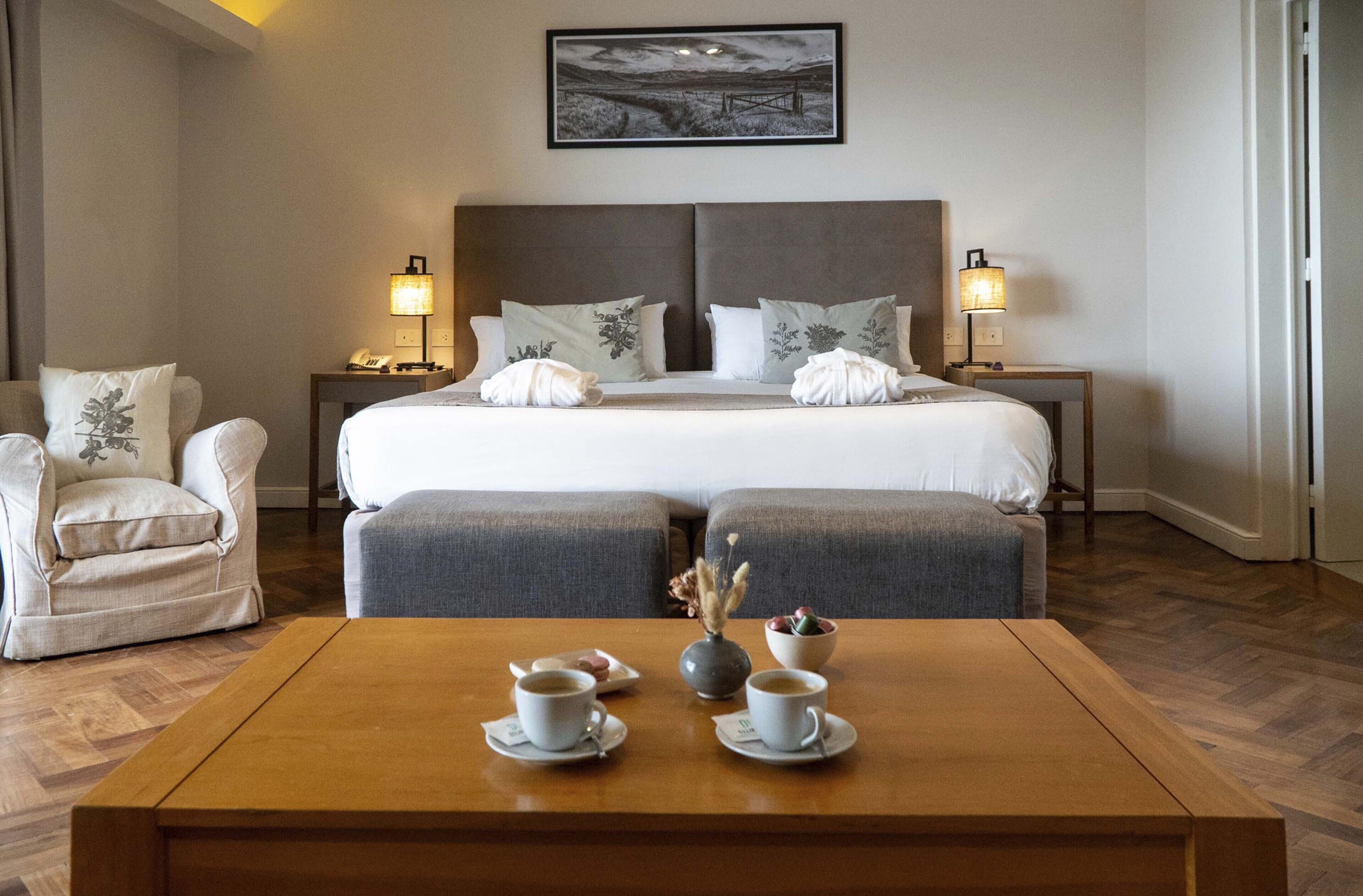 A hotel room with a large bed, two bedside tables with lamps, a chair, a coffee table with cups and a vase, and a painting on the wall.