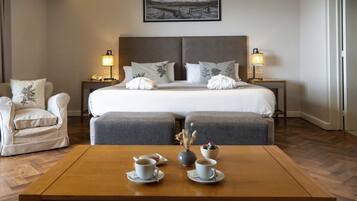 A hotel room with a large bed, two bedside tables with lamps, a chair, a coffee table with cups and a vase, and a painting on the wall.