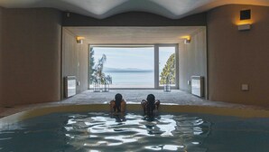 An indoor swimming pool with two people in the water, a view of the sea through large windows, and a ceiling with a recessed light. - La Cascada Casa Patagónica by DON (San Carlos de Bariloche)