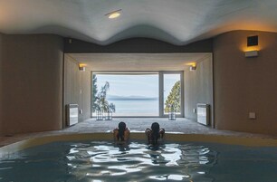 Una piscina interior con dos personas en el agua, vistas al mar a través de amplias ventanas y un techo con luces empotradas.