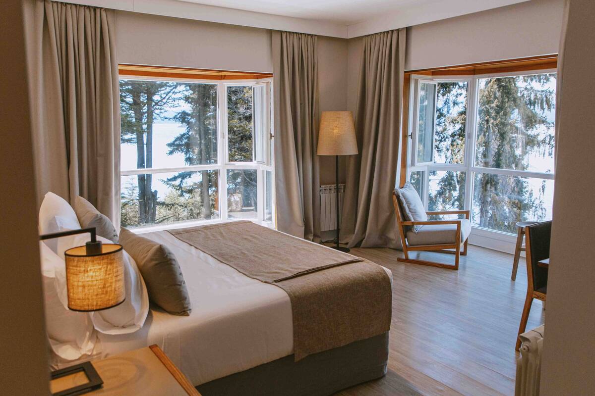 Ein Hotelzimmer mit Bett, Stuhl, Lampe und großen Fenstern mit Blick auf Bäume.