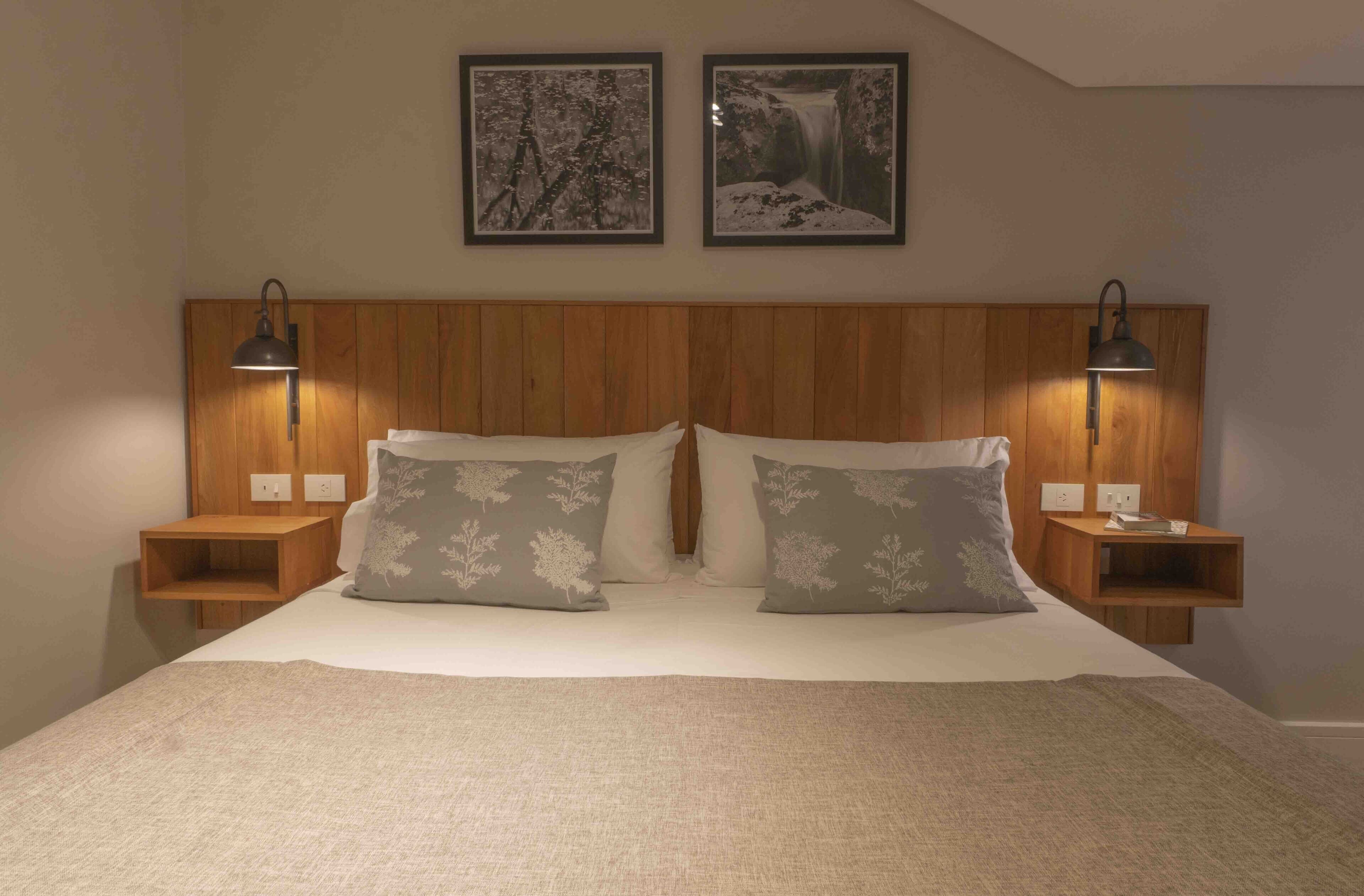 A bedroom with a wooden headboard, two bedside tables with lamps, a bed with a white comforter and two pillows with floral patterns, and two framed pictures on the wall.