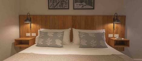 A bedroom with a wooden headboard, two bedside tables with lamps, a bed with a white comforter and two pillows with floral patterns, and two framed pictures on the wall.