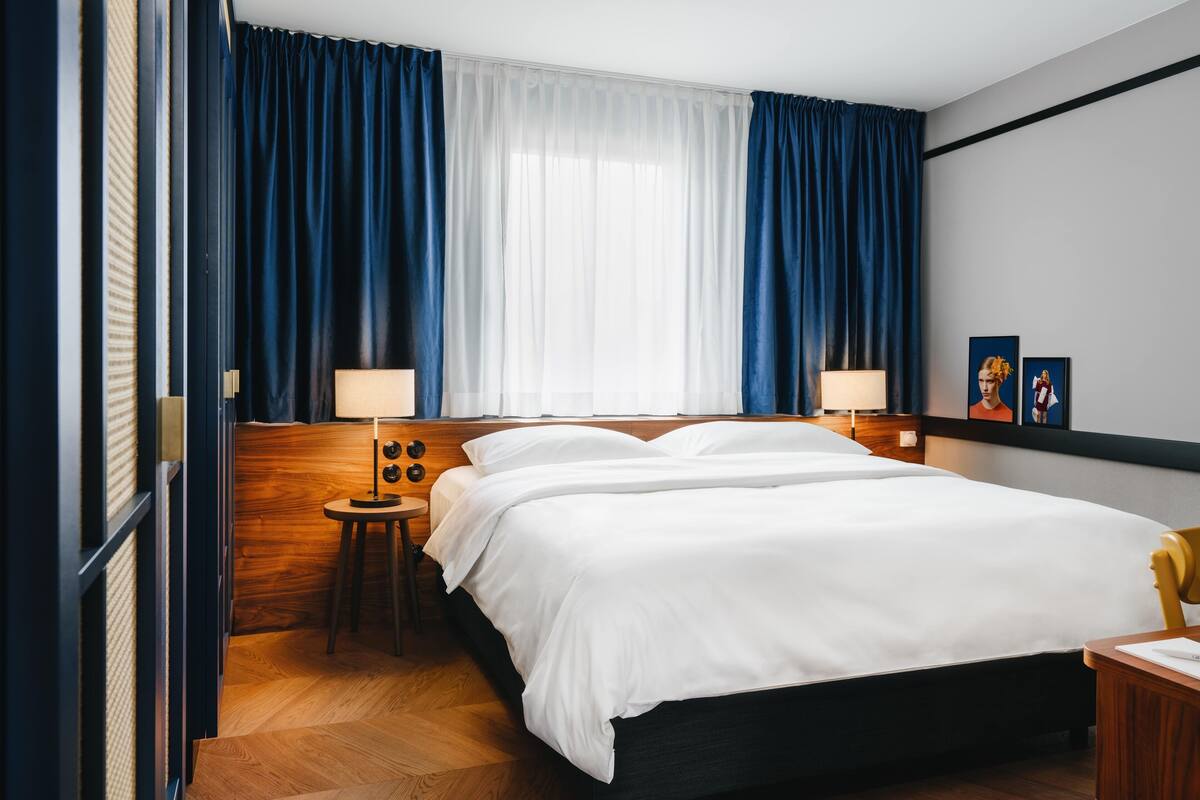Een hotelkamer met een groot bed, twee nachtkastjes met lampen, een houten hoofdeinde en een groot raam met blauwe gordijnen.