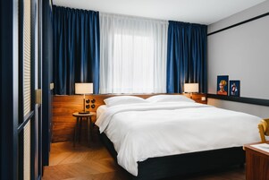 Quarto de hotel com uma cama grande, duas mesas de cabeceira com luminárias, uma cabeceira de madeira e uma janela ampla com cortinas azuis.