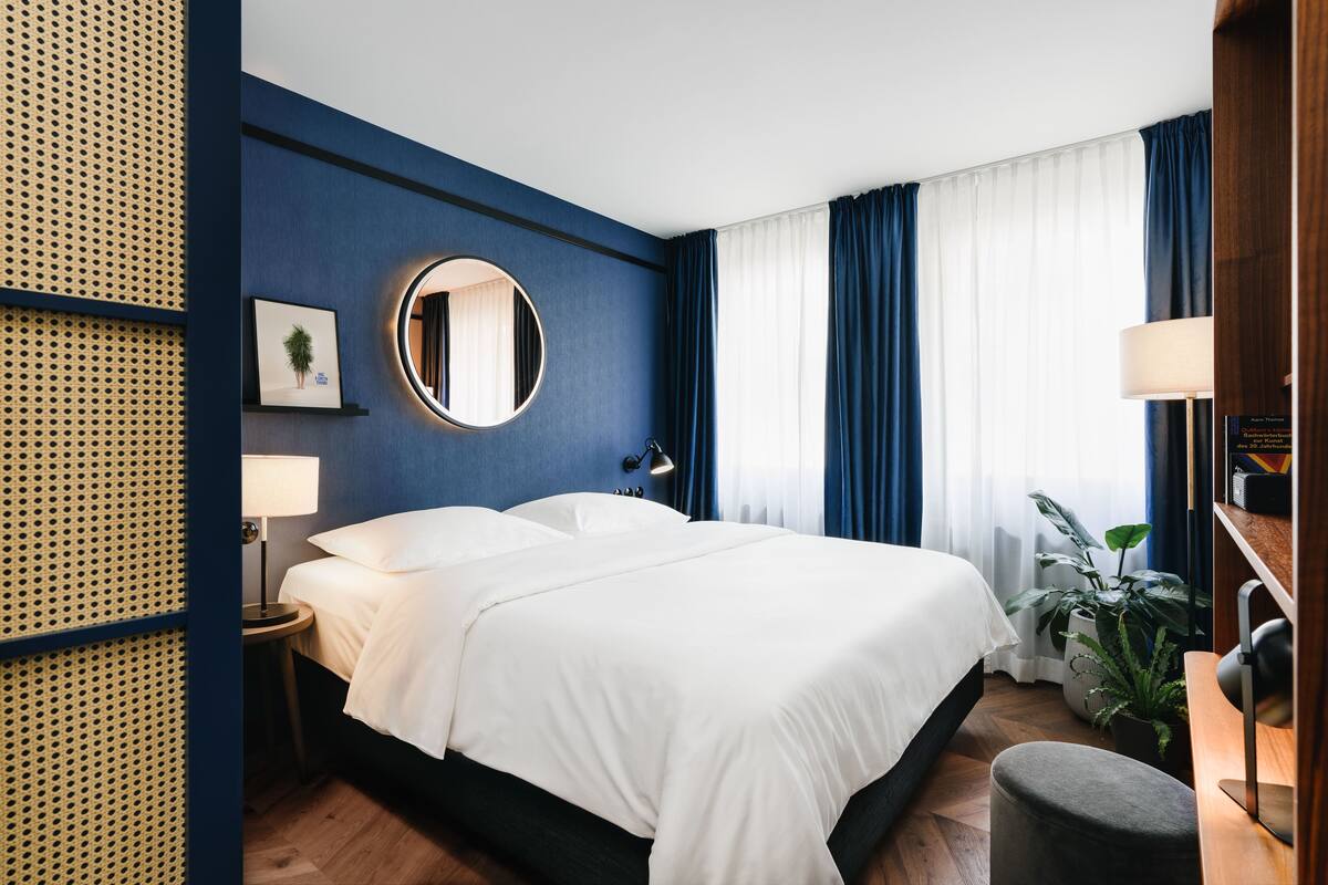 Een moderne hotelkamer met een groot bed, een ronde spiegel, een nachtkastje met lamp, een klein krukje en een potplant.