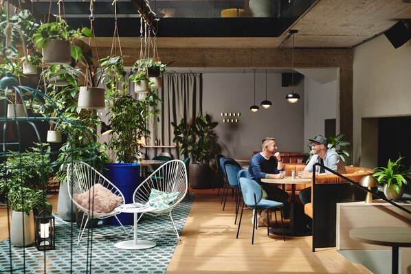 Um espaço interior moderno com plantas penduradas, diversos assentos incluindo uma cadeira branca e um sofá, e uma área de refeições com duas pessoas sentadas à mesa.