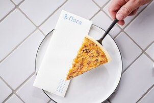 Uma fatia de pizza em um prato com uma guardanapo rotulado “& flora” e uma mão segurando um garfo.
