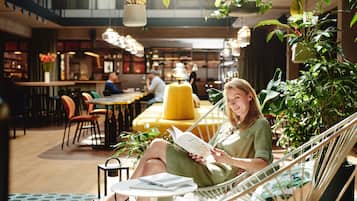 Una mujer leyendo un libro en un área de descanso con una silla hamaca, mesas, sillas y plantas colgantes.