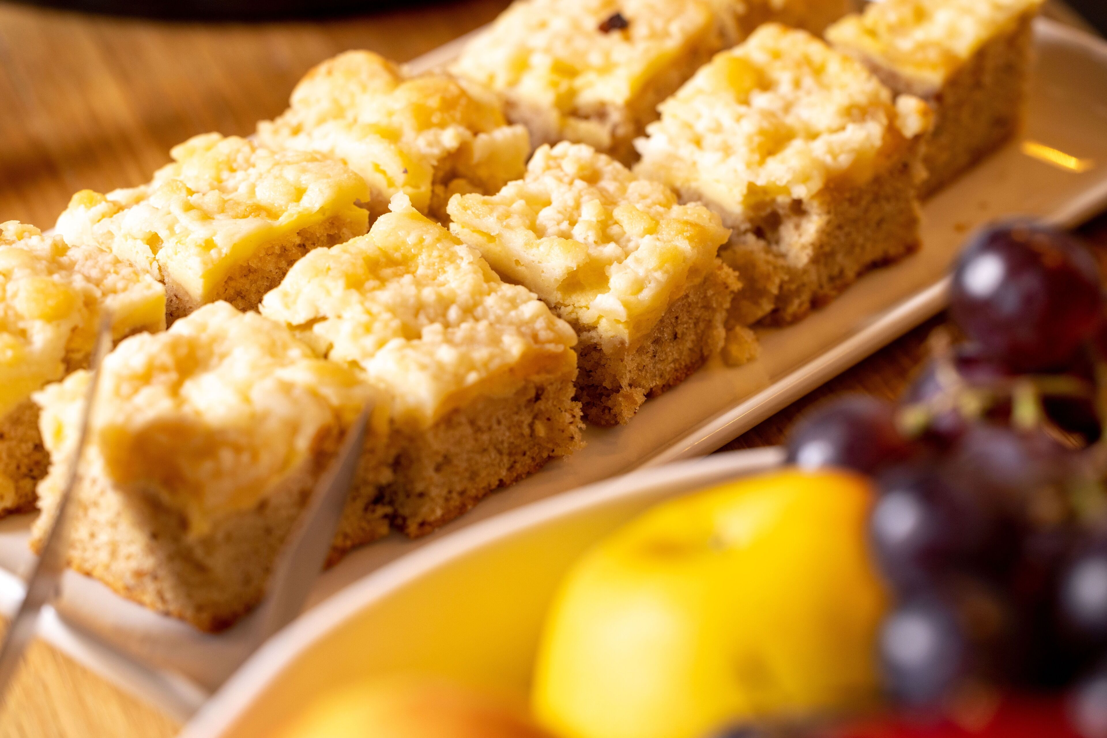 A tray of square-shaped desserts with a crumbly topping, accompanied by a bowl of grapes and a lemon.