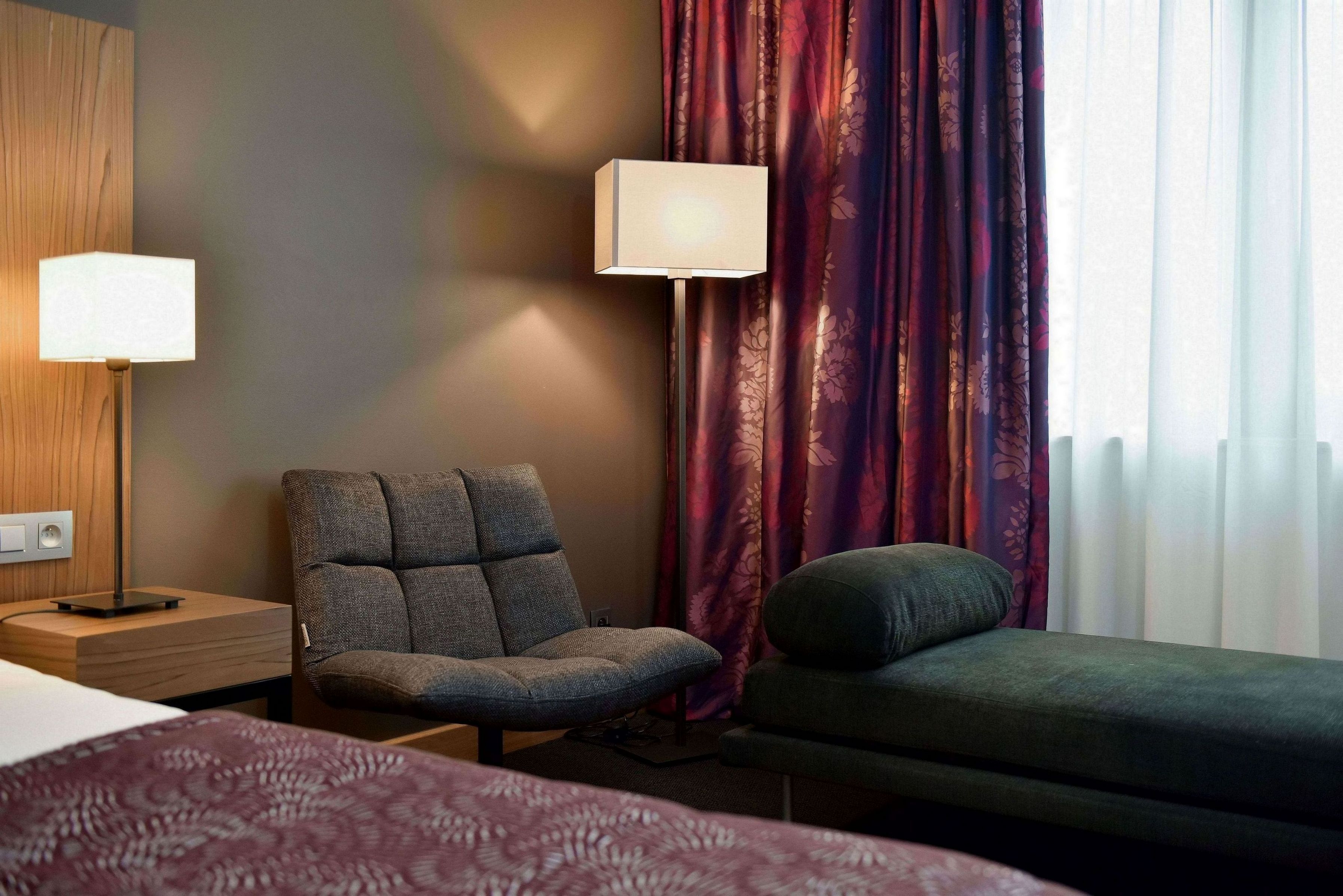 Une chambre d’hôtel avec un lit, une chaise, un canapé, une lampe et une fenêtre avec des rideaux.