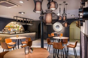 Zona de bar moderna con mesas y sillas redondas, barra con gran variedad de botellas y lámparas colgantes originales.