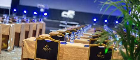 Un salón de conferencias con filas de sillas y mesas con la marca Martin's Hotels, equipado con botellas de agua y una pantalla de proyector al fondo.