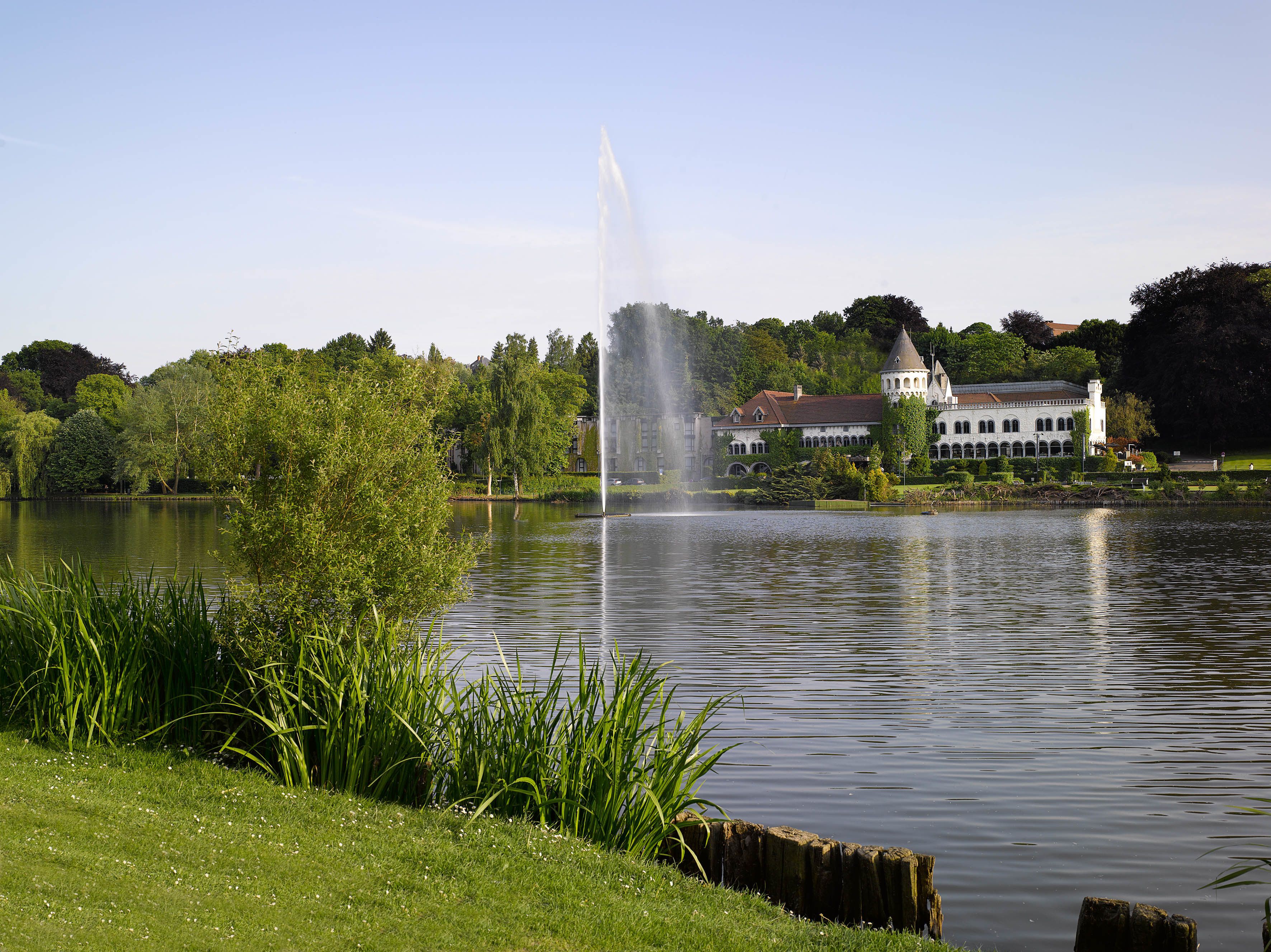 A serene lake with a fountain in the center, surrounded by lush greenery and a historic building with a tower.