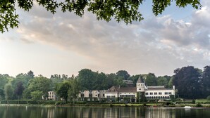 A large, historic building with multiple wings and a central tower, situated by a calm lake with reflections.