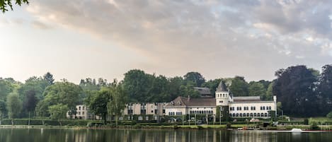 Un edificio grande e histórico con varias alas y una torre central, ubicado junto a un lago tranquilo con reflejos.