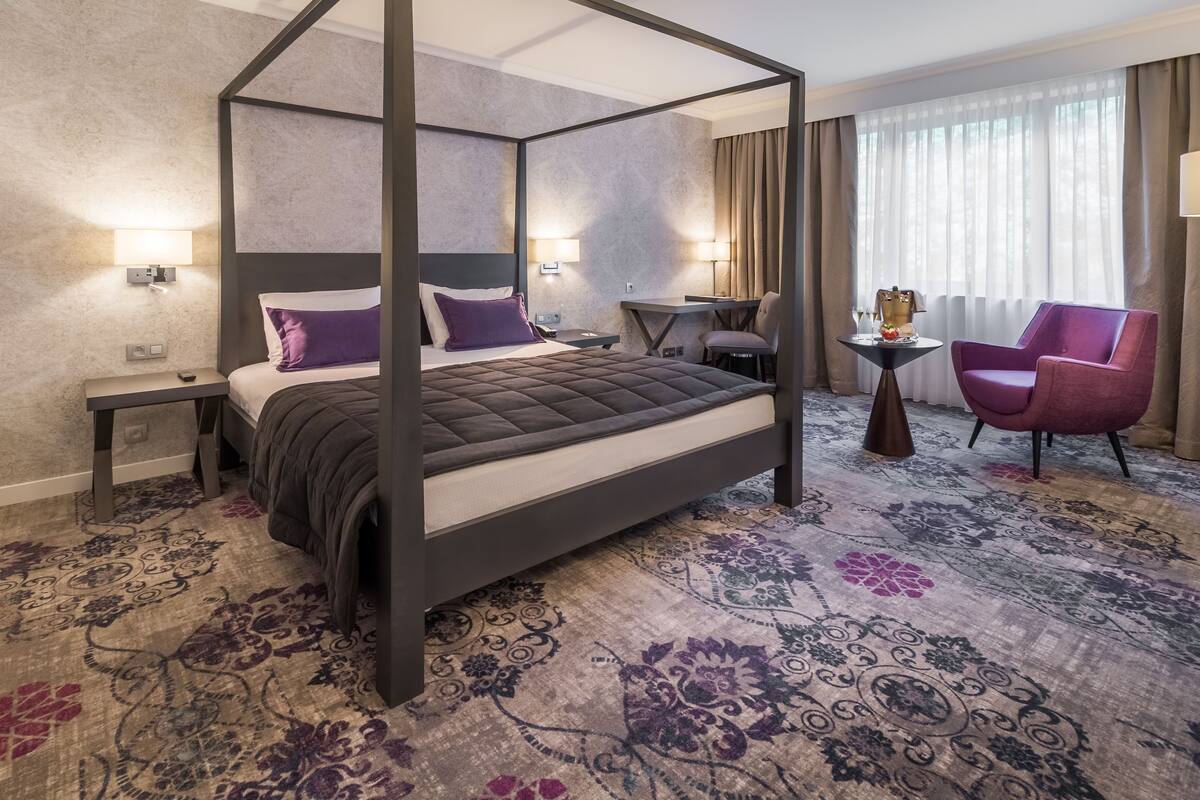 Uma cama de dossel com cabeceira escura, uma escrivaninha com cadeira e uma poltrona roxa num quarto de hotel com carpete estampado.