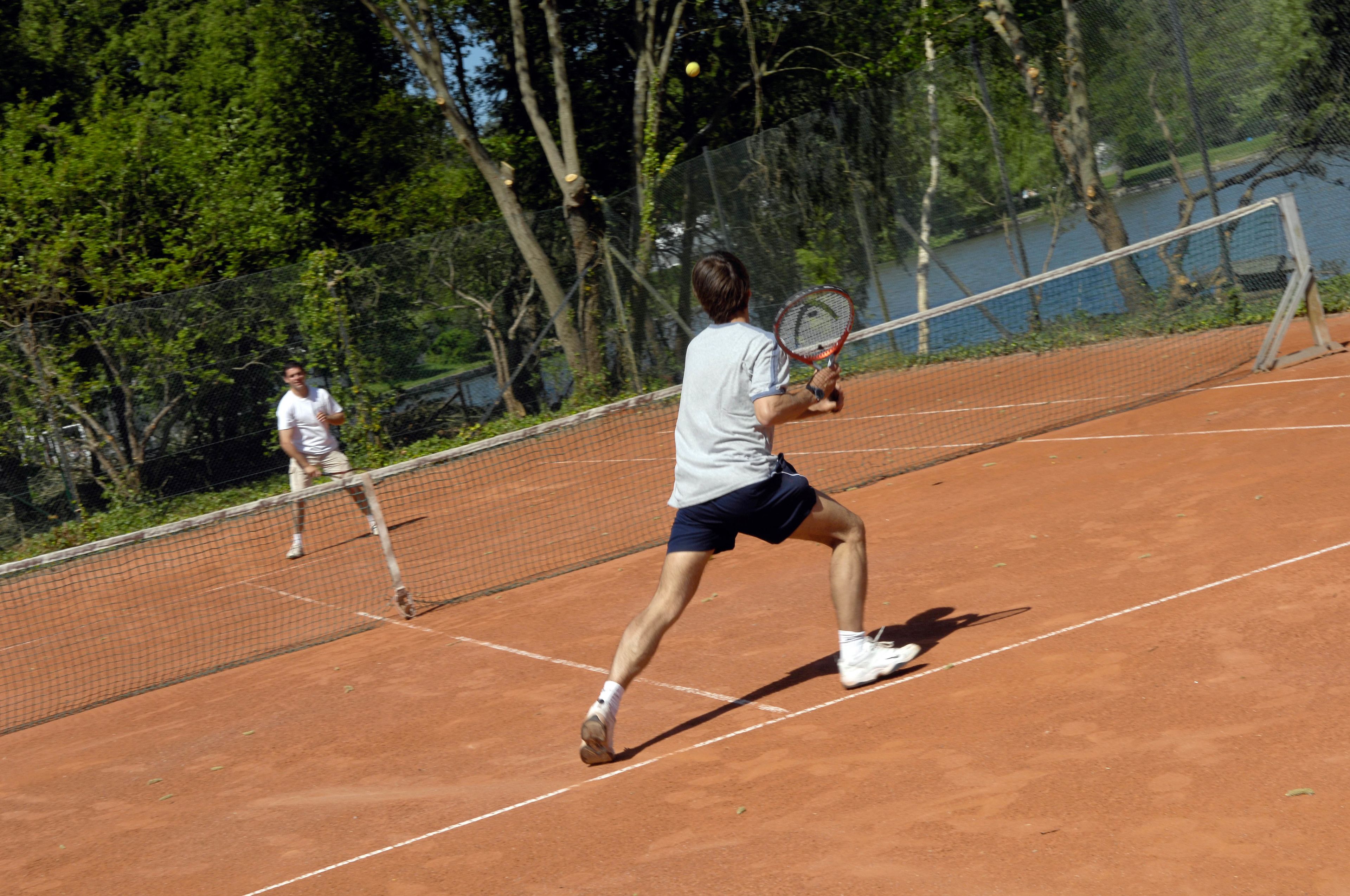 Ein Tennisplatz mit Netz, umgeben von Bäumen und einem Gewässer.