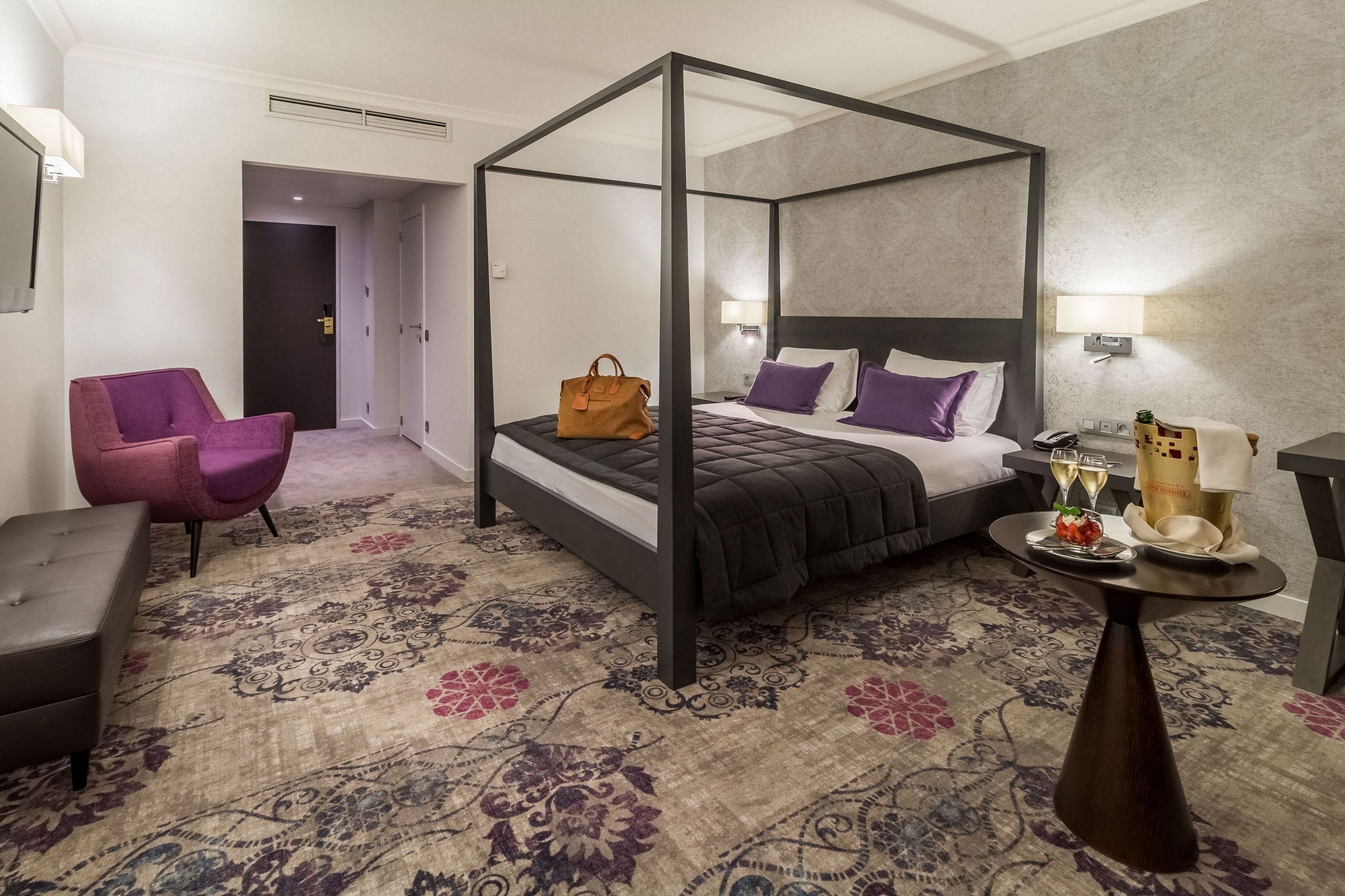 A four-poster bed with a dark headboard, a purple armchair, a small round table with a bottle and glasses, and a patterned carpet.