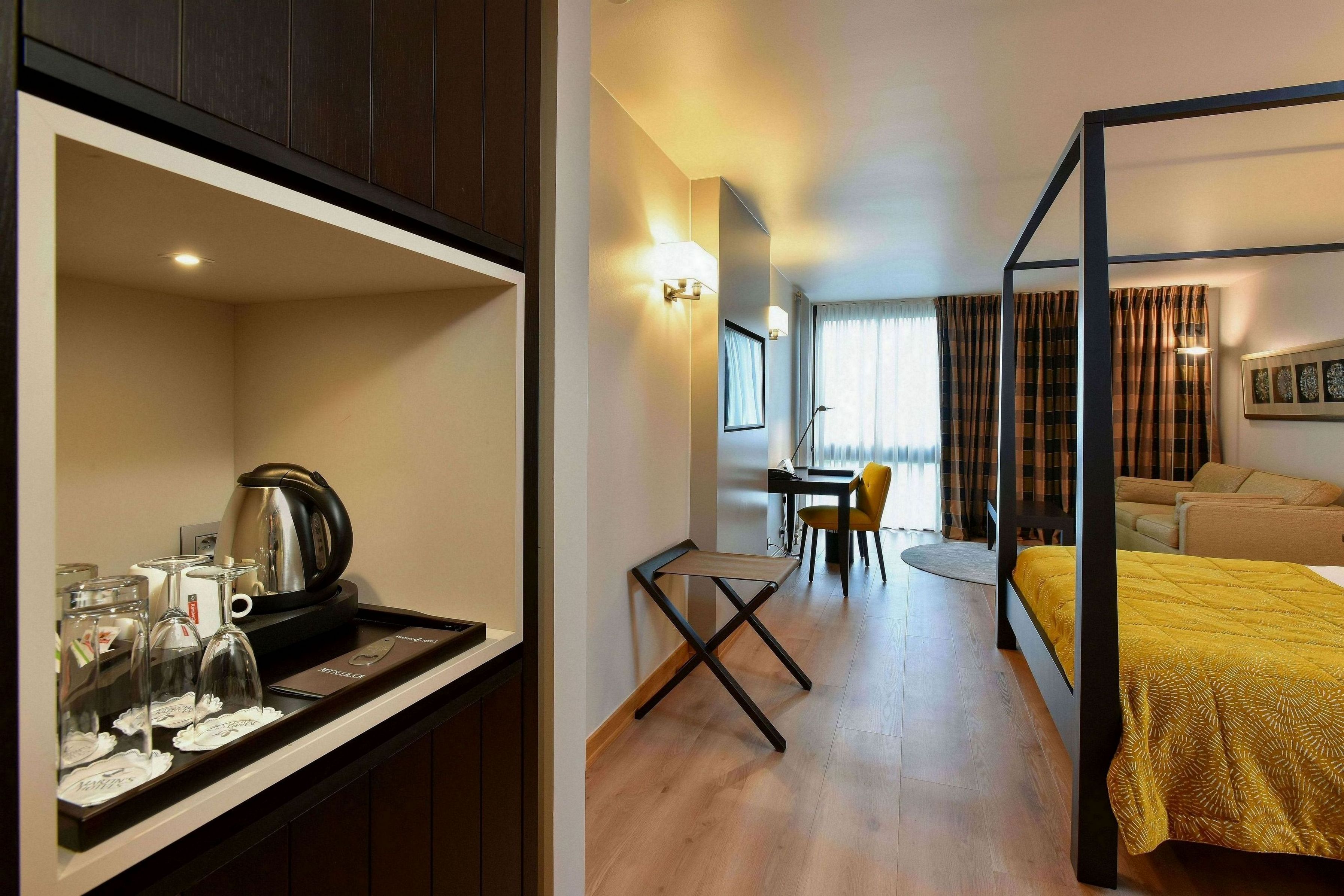 A hotel room with a bed, a desk, and a chair. There is a coffee maker on the desk.