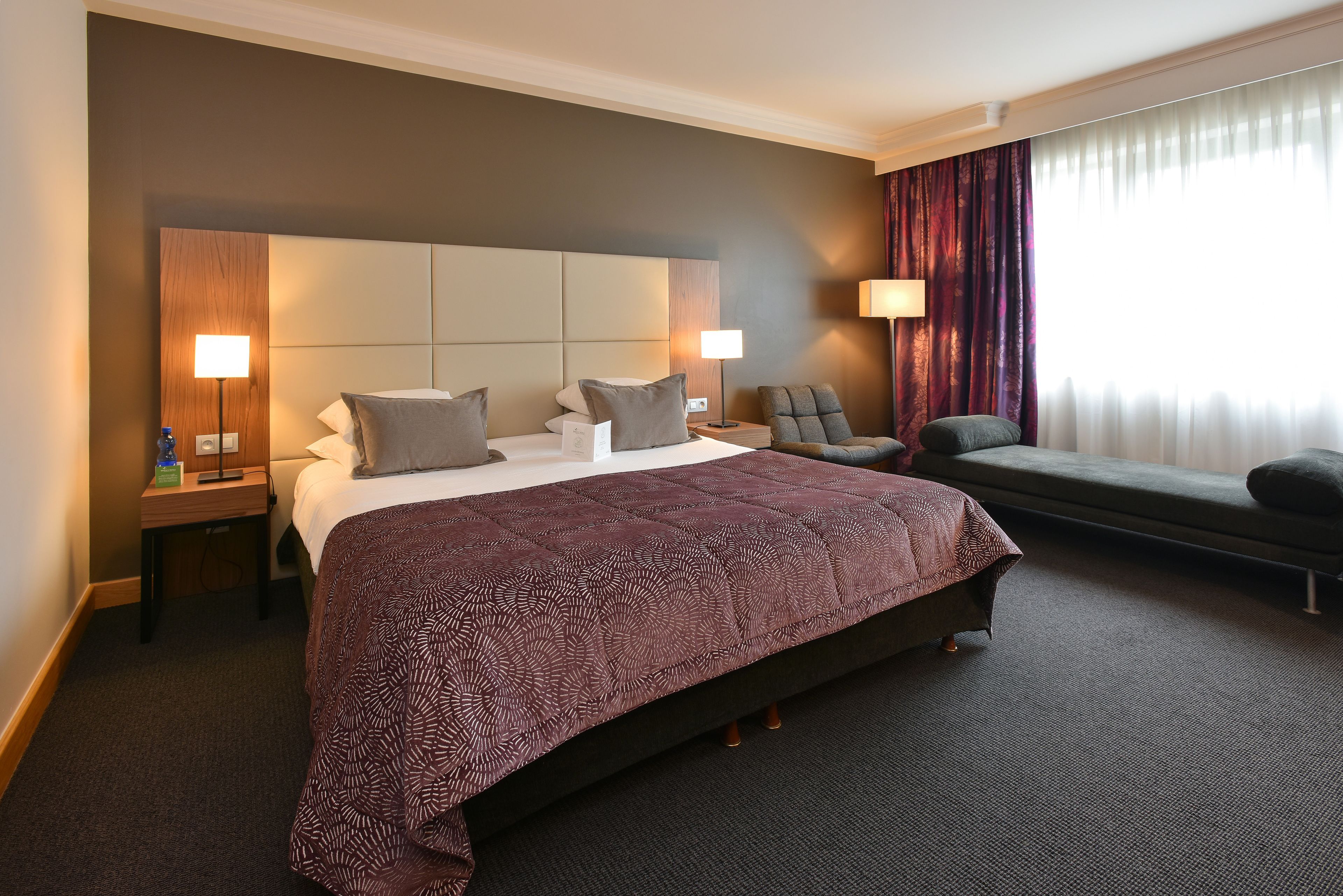 A hotel room with a large bed, two bedside tables, a chair, a sofa, and a window with curtains.