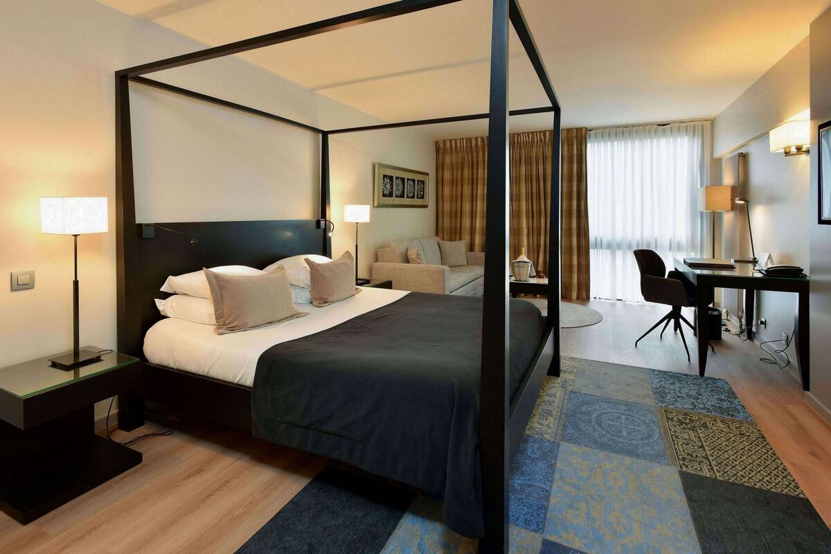 Uma cama de dossel com edredom escuro, uma escrivaninha com cadeira, luminária e janela com cortinas.