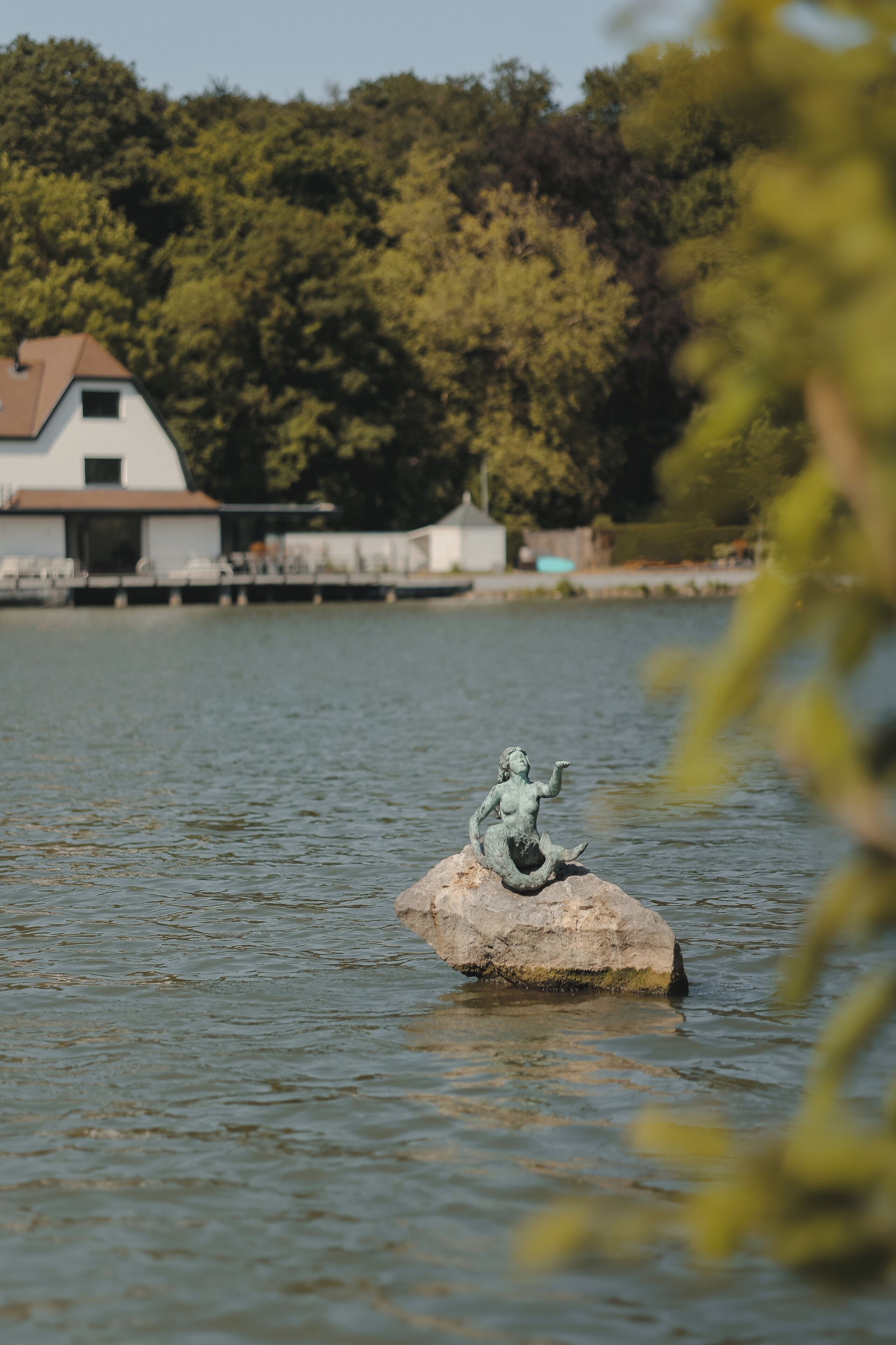 Una estatua sobre una roca en el agua, con un edificio y árboles al fondo.