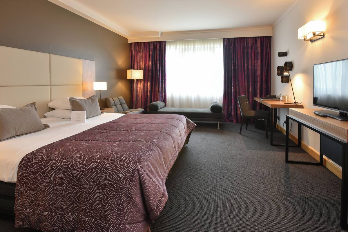 Quarto de hotel com uma cama grande, uma escrivaninha, uma cadeira, uma televisão e uma janela com cortinas.