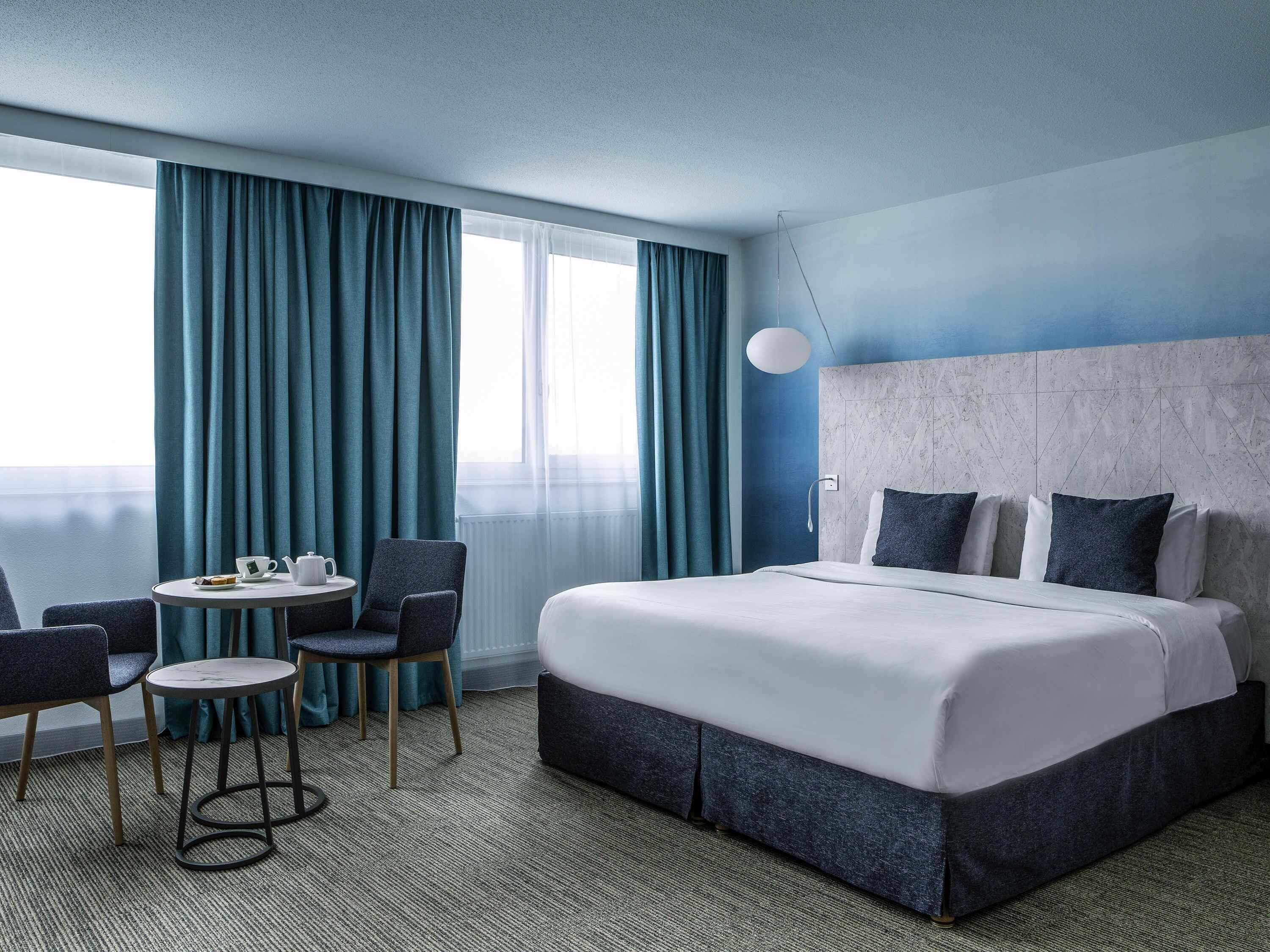 A hotel room with a bed, a small table with chairs, a view of the sea, and a large window with curtains.