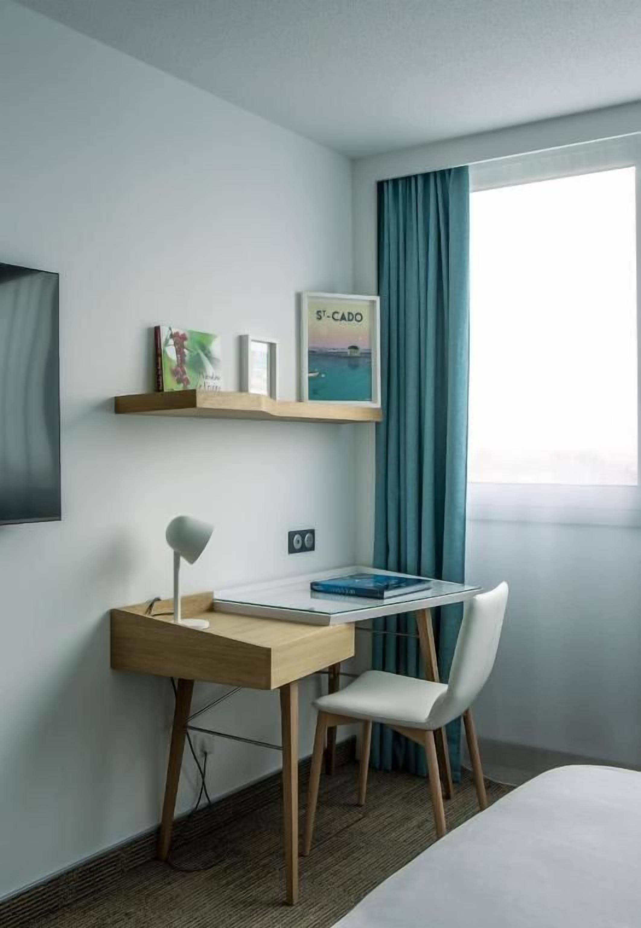 A hotel room with a bed, a desk with a chair, a shelf with books and a picture, and a window with curtains.