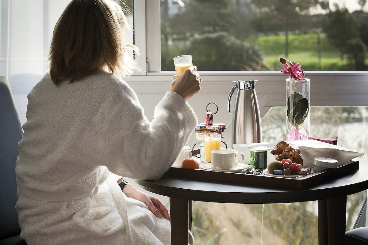 A person in a white robe sitting by a window with a tray of breakfast items, including a croissant, fruit, and a glass of orange juice.