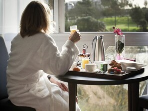 A person in a white robe sitting by a window with a tray of breakfast items, including a croissant, fruit, and a glass of orange juice. - Mercure Vannes Le Port (Vannes)