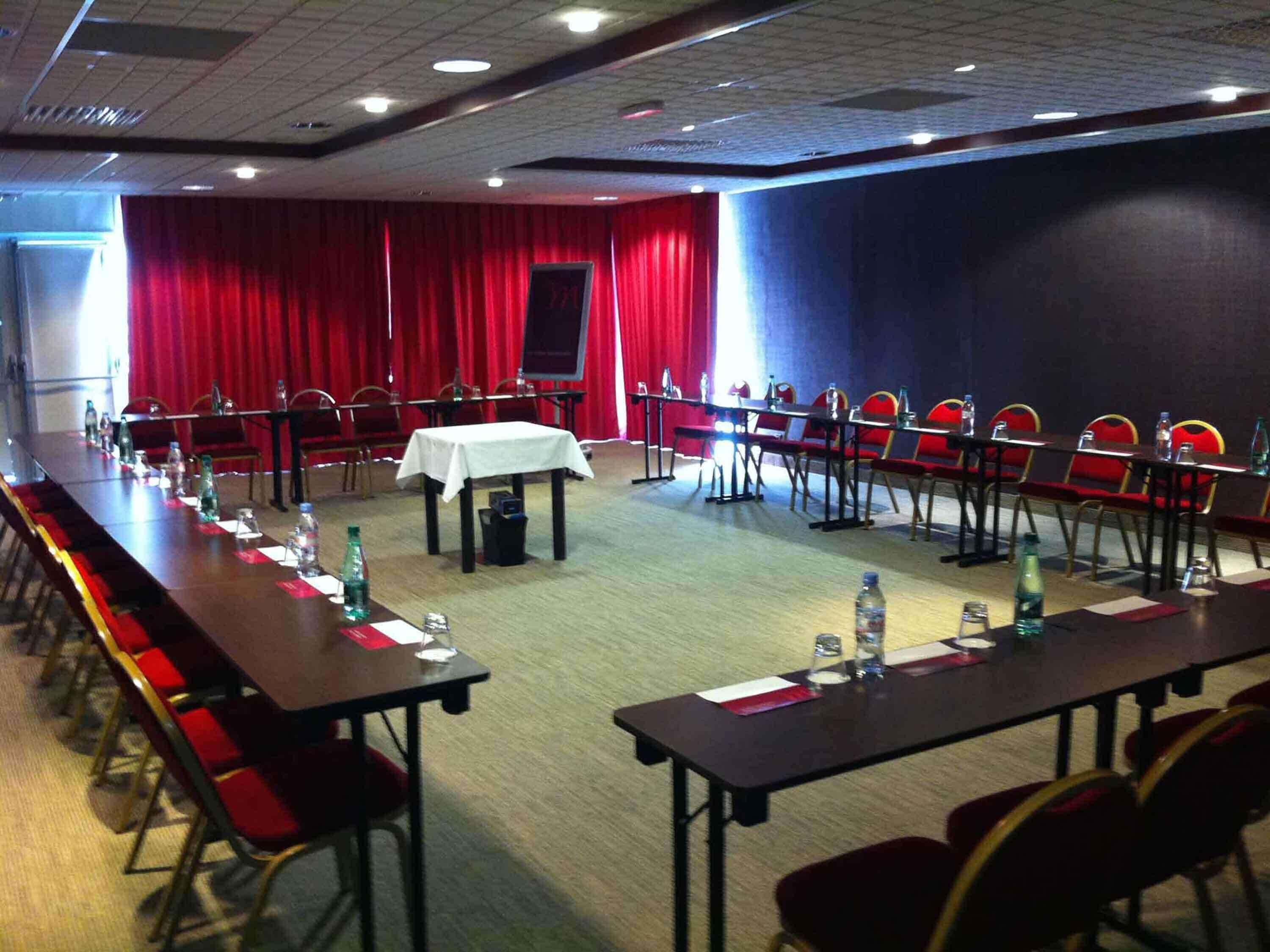 A conference room with long tables, red chairs, and a stage with a red curtain.