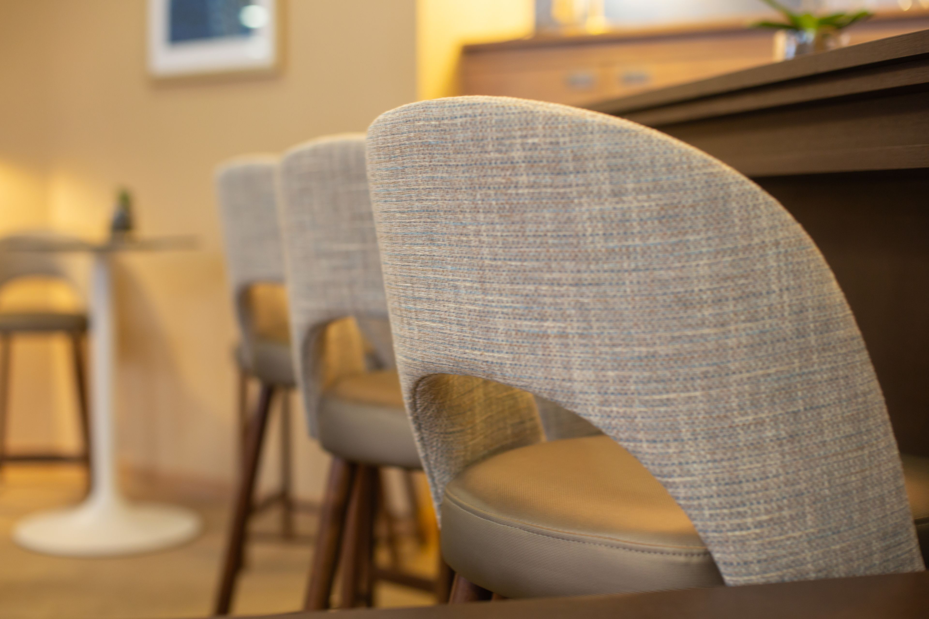A row of upholstered chairs with a woven texture, arranged in a dining area with a wooden table and a bar counter in the background.