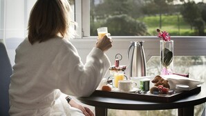 Una persona vestida con una bata blanca sentada junto a una ventana con una bandeja de desayuno, que incluye una cafetera, un vaso de jugo de naranja y un florero con flores.