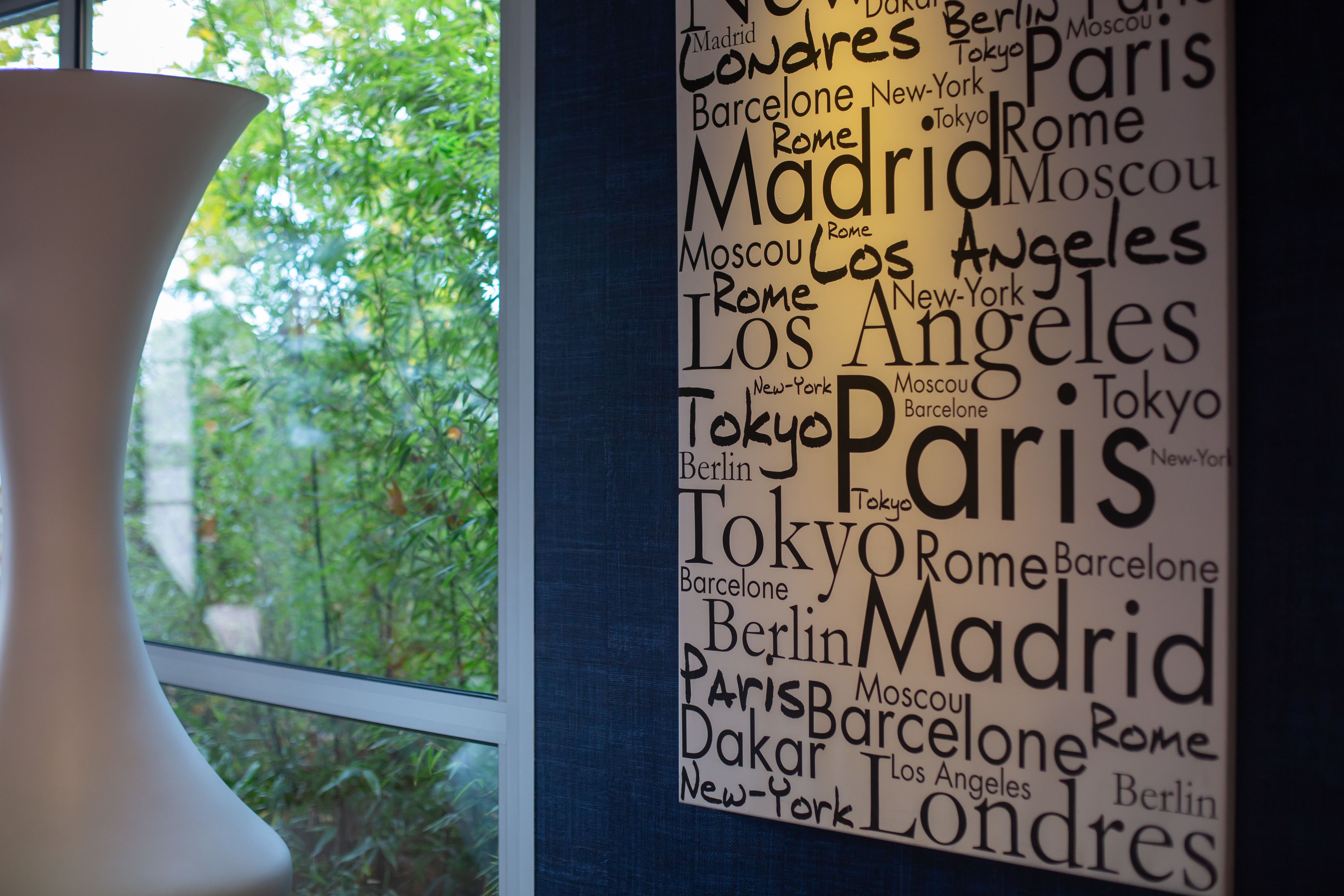 A wall with a word cloud of city names, a window, and a white vase.