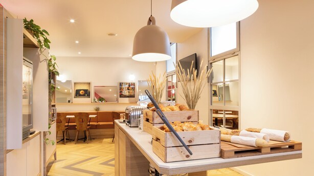 A modern kitchen with wooden cabinets, a built-in oven, and a countertop with bread and pastries. - ibis Styles Paris Nation Porte De Montreuil (Paris)