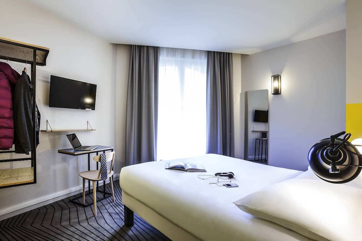 A hotel room with a bed, a desk with a laptop, a chair, a TV, and a coat rack.