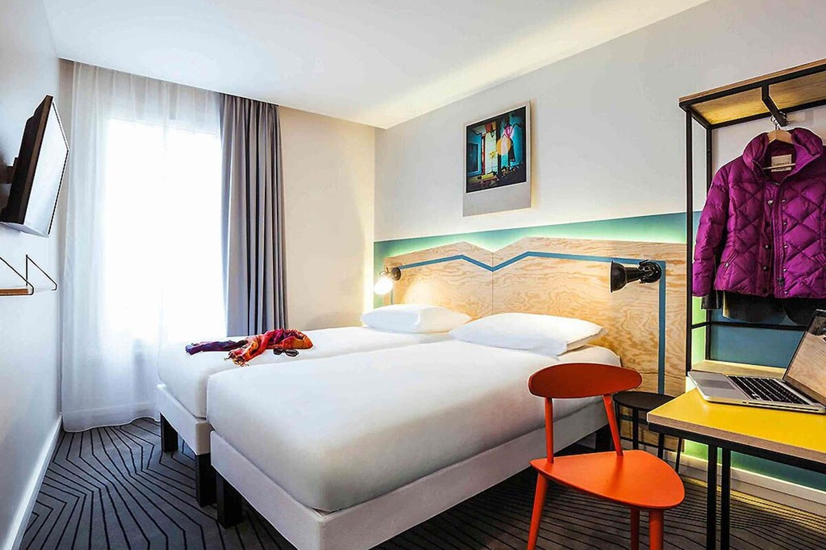 A hotel room with a bed, a desk with a laptop, a chair, a coat rack with a jacket, and a painting on the wall.