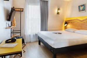 A hotel room with a bed, a desk with headphones and a book, a TV, and a window with curtains. - ibis Styles Paris Nation Porte De Montreuil (Paris)