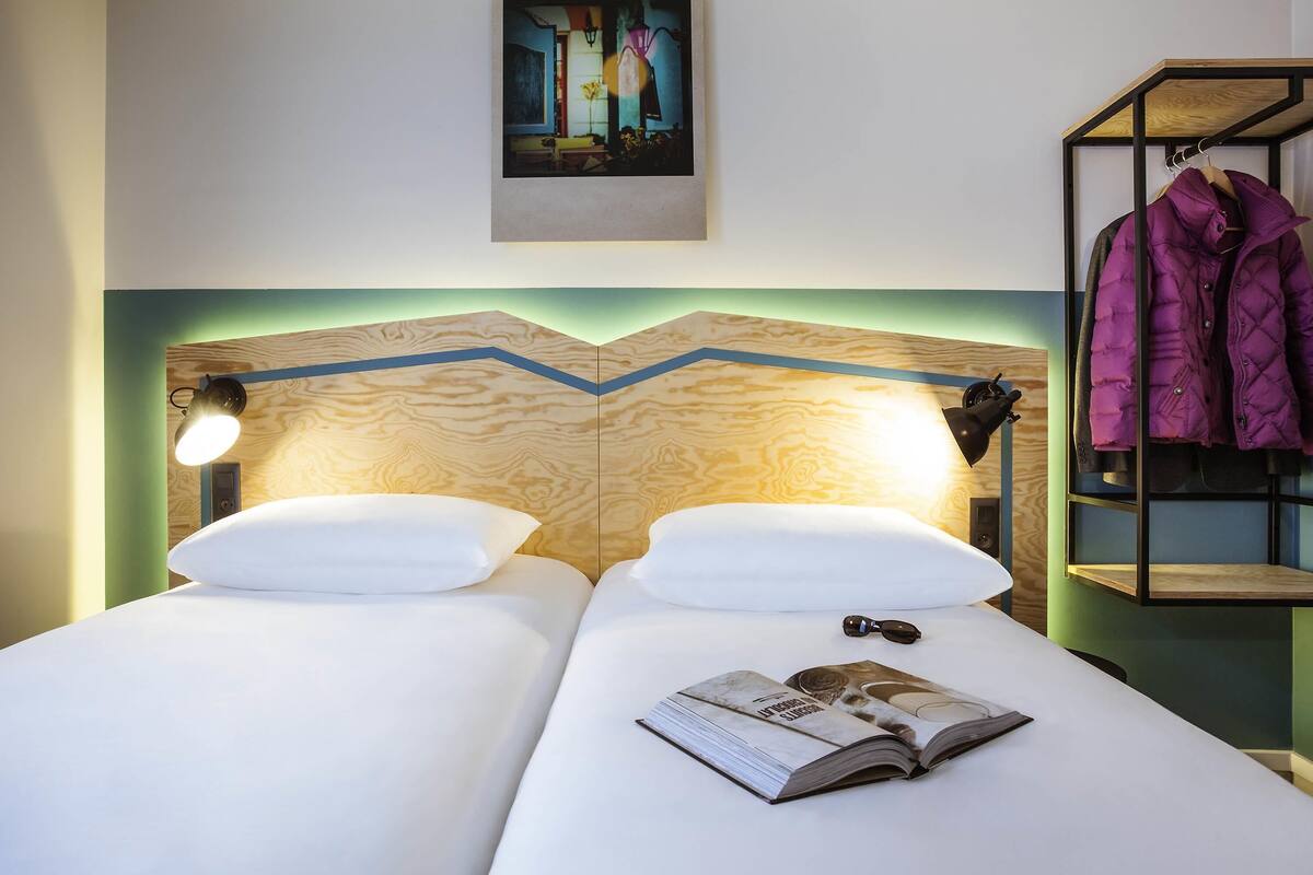 A double bed with white linens, a wooden headboard with a blue accent, a framed picture above the bed, and a coat rack with a purple jacket.