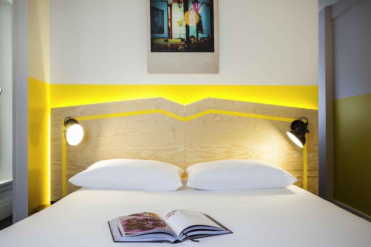 A neatly made bed with two pillows, a book on the bed, and two wall-mounted lamps.